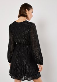 Czarna sukienka z teksturowanym wzorem w zygzak, długimi bufkami i dopasowanym stanem. Przezroczysty materiał z subtelnym połyskiem, długość do kolan.