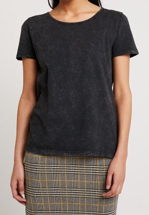 Frau trägt ein ausgebleichtes schwarzes kurzärmliges Shirt und einen hoch taillierten karierten Rock mit beigen, schwarzen und weißen Mustern.