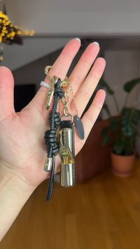 Portachiavi con cilindro in metallo dorato lucido, cinturino in pelle nera e dettagli decorativi. Presenta catene e un'etichetta scura.