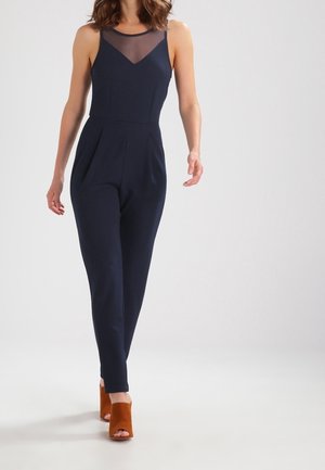 Granatowy kombinezon z przezroczystym siateczkowym dekoltem, dopasowaną talią i zwężającymi się nogawkami. W połączeniu z brązowymi sandałami na obcasie z otwartym przodem.