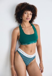 Grön bomullssportbh med en bred band som har vit "Calvin Klein" -text, kombinerad med mönstrade teal trosor med vit midjeband.