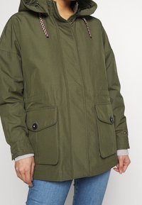 Una persona che indossa una giacca a vento verde oliva con tasche a pattina frontali e dettagli in bottone, abbinata a jeans blu.