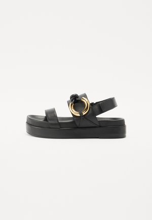 Sandal de couro preto com uma plataforma grossa, apresentando duas tiras e um detalhe circular dourado. Superfície texturizada e tira ajustável para o tornozelo.