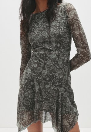 Woman wearing a long-sleeve, dark paisley-patterned sheer dress with an asymmetrical hem, standing against a plain light background.