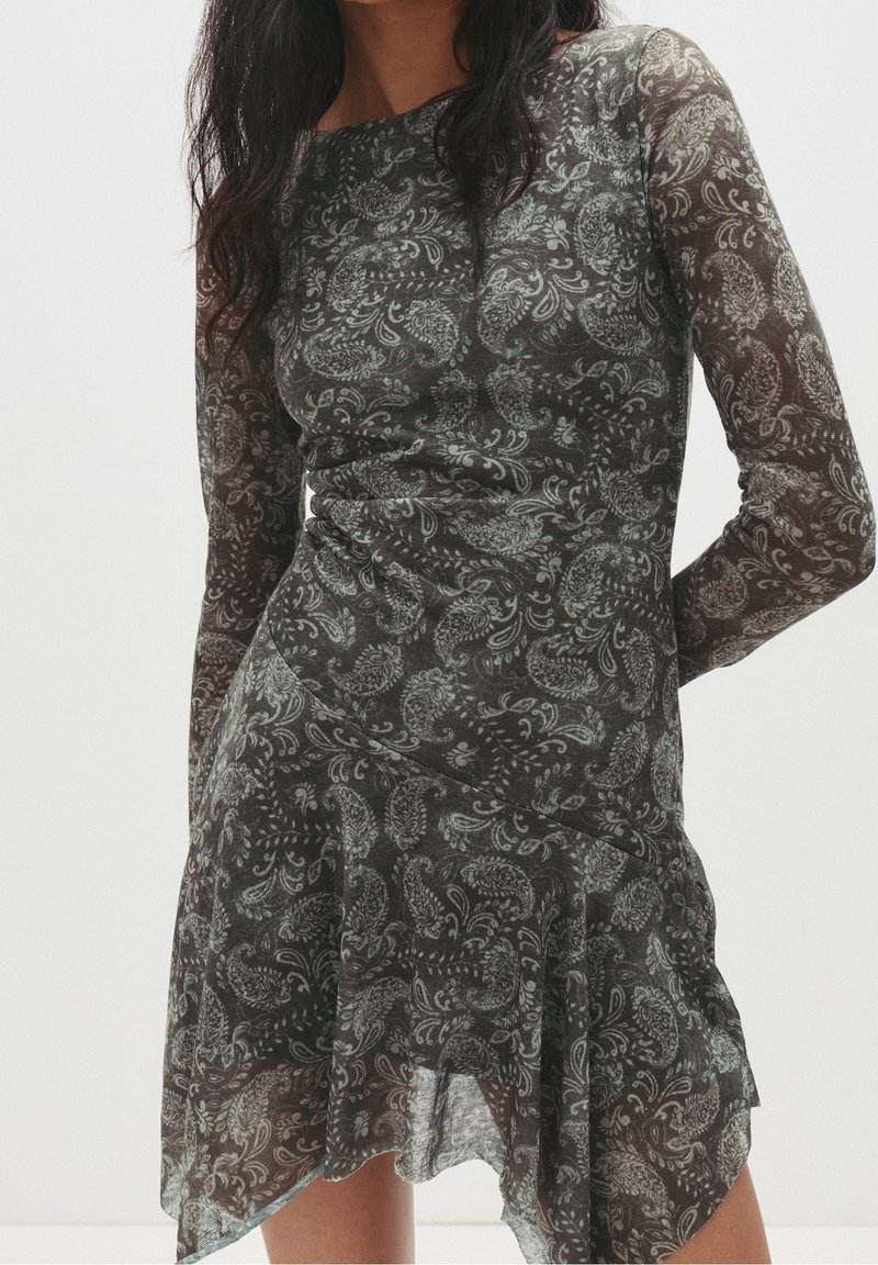 Femme portant une robe longue transparente à manches longues, motif paisley foncé, avec un ourlet asymétrique, debout devant un fond clair uni.