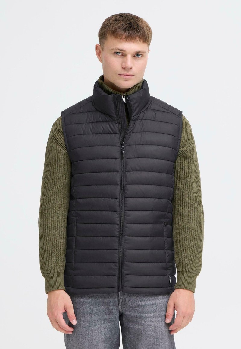 Svart polstret vest med høy krage, med horisontal quilt, frontglidelås og to sidelommer. Brukt over en grønn ribbestrikket genser.