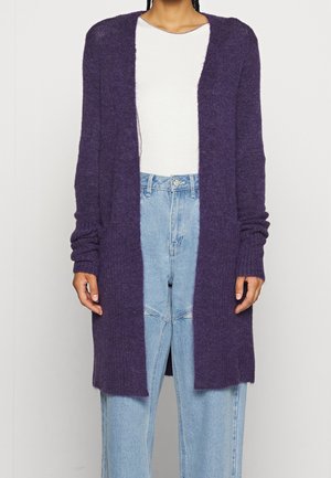 Person iført en lang lilla åben-front cardigan, hvid skjorte og lyseblå højtaljede jeans mod en ensfarvet baggrund.