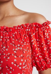 Kvinders skulder iført en rød off-shoulder top med elastisk halsudskæring og hvidt blomstermønster på kort pufærme.