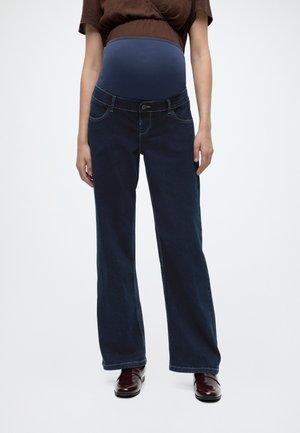 Gravid person iført mørkeblå graviditetsjeans med elastisk panel og brun kortærmet top, stående på hvid baggrund.