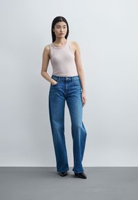 Jeune femme debout, une main dans la poche, portant un haut sans manches rose clair, un jean bleu à jambes larges et des chaussures noires pointues.