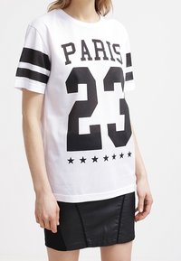 Person som bär en vit sportig skjorta med "PARIS 23" och svarta stjärnor, i kombination med en svart läderkjol.