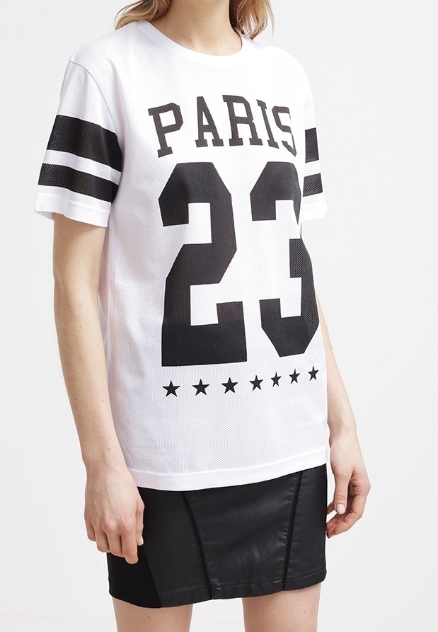 Person som bär en vit sportig skjorta med "PARIS 23" och svarta stjärnor, i kombination med en svart läderkjol.