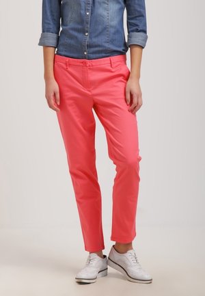 Femme portant un pantalon slim rose corail, une chemise en denim retroussée et des chaussures brogues blanches, debout devant un fond uni.