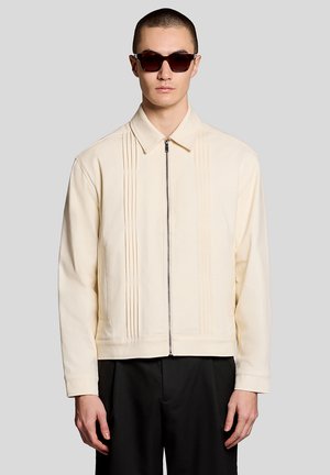 Homme portant des lunettes de soleil noires, une veste crème zippée avec des coutures verticales, et un pantalon noir, sur fond gris uni.