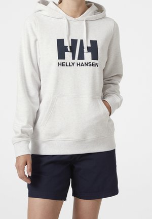 Personne portant un sweat à capuche Helly Hansen gris clair avec une poche avant et un short bleu marine foncé, debout avec une main dans la poche.