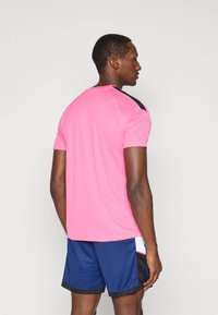 Αθλητική μπλούζα εφαρμοστής γραμμής σε φωτεινό ροζ με μαύρες λεπτομέρειες στους ώμους, συνδυασμένη με ναυτικές βερμούδες που διαθέτουν σχέδιο πλαϊνής πάνελ και μαύρη περίμετρο.