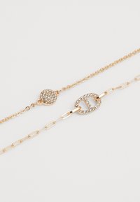 Gold-tone layered bracelet featuring a crystal-studded link and round charm, with chain and oval hardware accents. Smooth texture throughout.