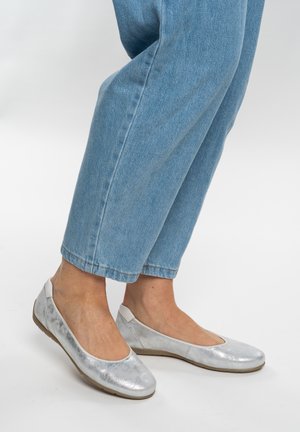 Beine in hellblauen Jeans, die silberne Ballettschuhe mit abgerundetem Zehenbereich und beigen Sohlen auf weißem Hintergrund tragen.