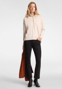 Pullover beige chiaro con chiusura a bottoni, maniche lunghe e polsini a coste; abbinato a jeans neri a zampa; stivali neri alla caviglia; modella che tiene un giubbotto.