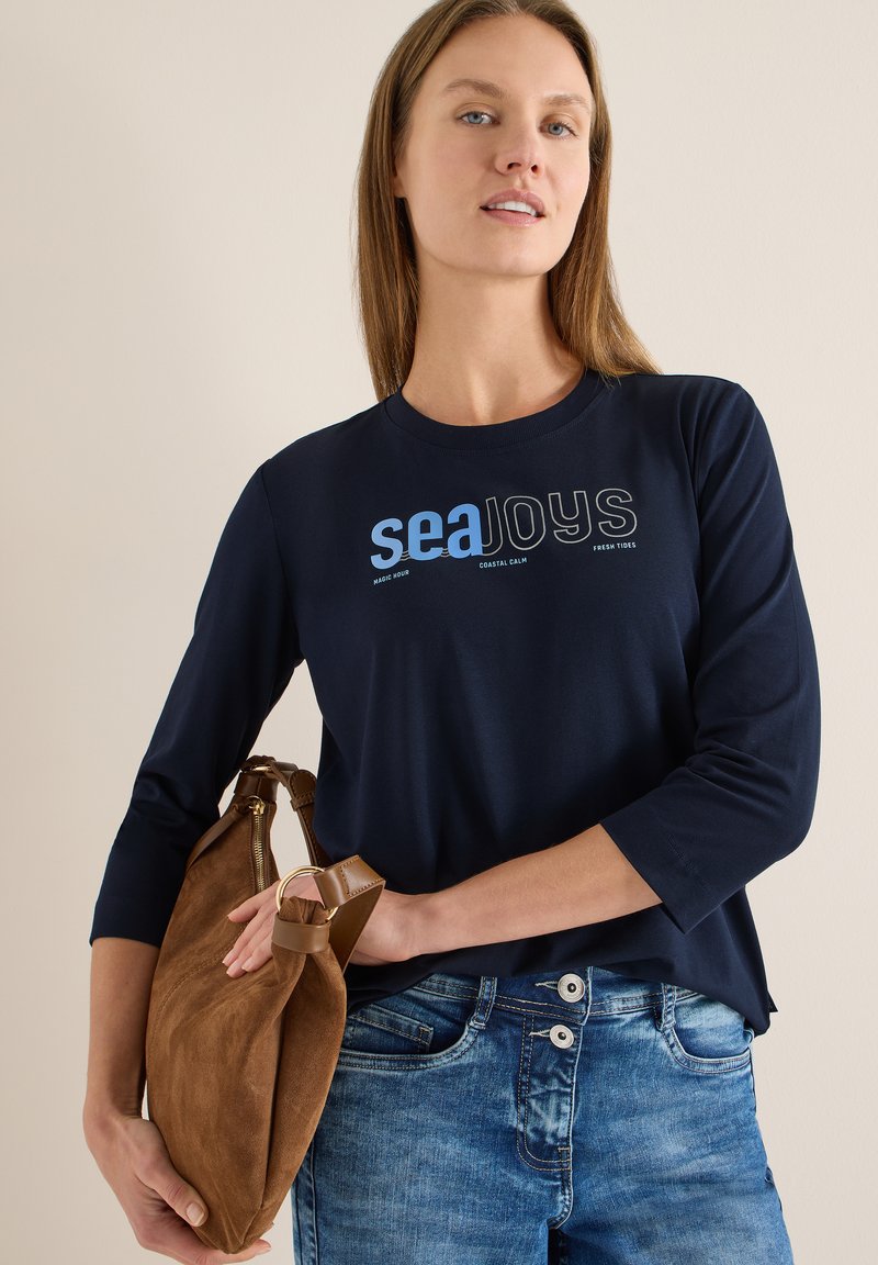 Chemise bleu marine à manches longues avec imprimé "seaJoys", associée à un jean bleu clair et un sac en suède marron avec des accents en cuir.
