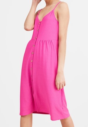 Femme portant une robe rose sans manches à boutons sur le devant, avec des bretelles fines et une taille froncée, debout devant un fond blanc uni.