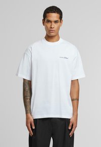 White cotton T-shirt with short sleeves and a crew neckline, featuring a small blue logo text on the left side. Smooth texture and relaxed fit.