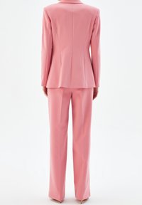 Jasnoróżowy garnitur z dopasowaną marynarką i szerokimi spodniami, wyróżniający się gładką teksturą i zorganizowanym krojem. Brak widocznych elementów metalowych.