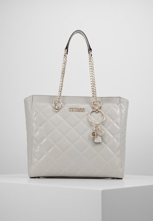 Sac cabas matelassé gris clair brillant avec poignées chaîne en or, logo GUESS et breloques dorées décoratives suspendues à l'avant.