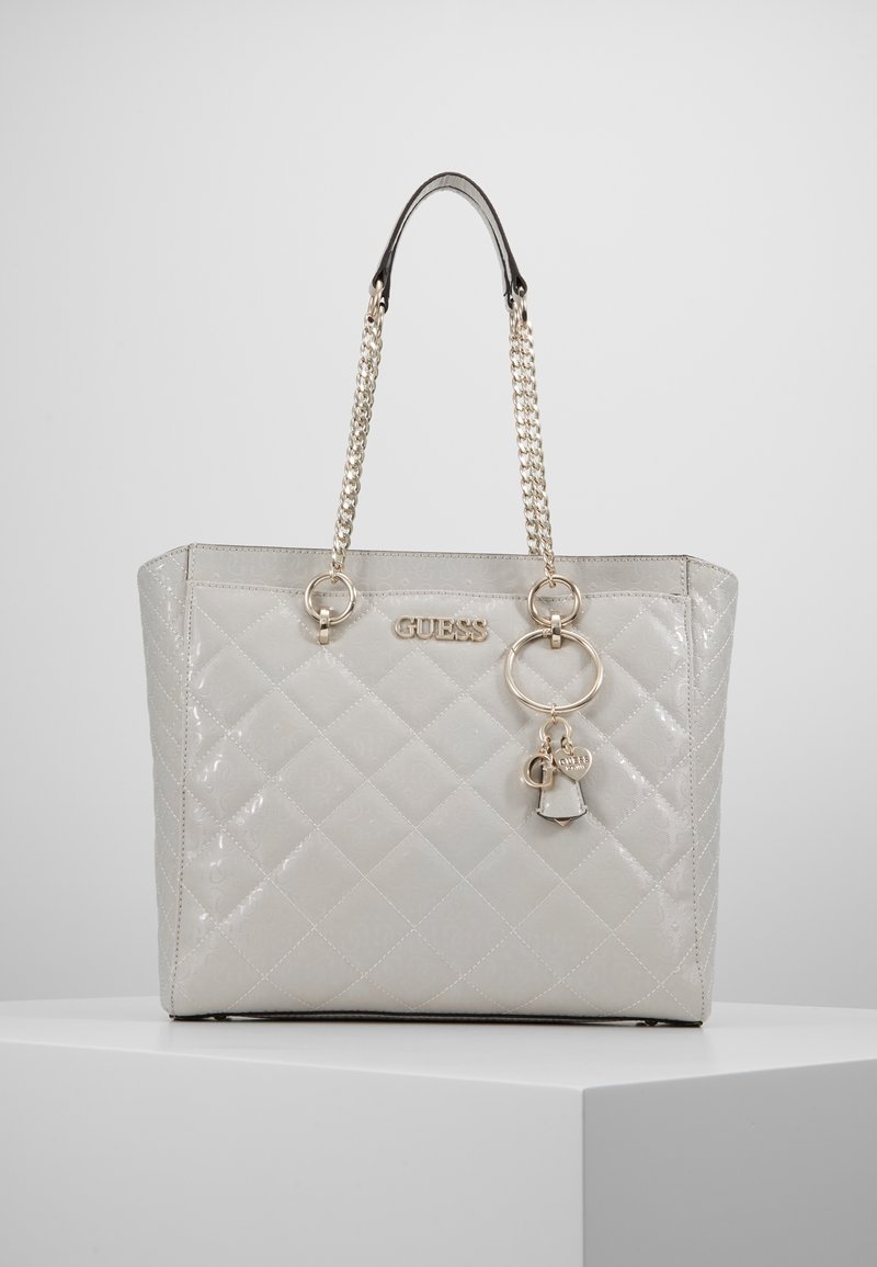 Sac cabas matelassé gris clair brillant avec poignées chaîne en or, logo GUESS et breloques dorées décoratives suspendues à l'avant.