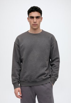 Un homme pose avec un sweat-shirt gris foncé à col rond et un pantalon assorti sur un fond uni et clair.