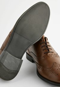Pantofi de piele maro, cu detalii brogue, având o talpă din cauciuc și o textură netedă. Pantofii au un design cu șireturi.