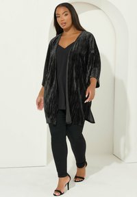 Zwarte, samengeperste fluwelen cardigan met een open voorkant, wijde mouwen en een knielange snit, gecombineerd met een zwarte top en slim-fit zwarte broek.