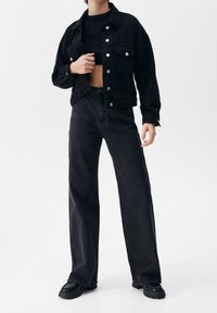 Zwarte spijkerjas met zilveren knopen, verkort ontwerp, gecombineerd met een high-waisted zwarte wijde pijpen jeans en chunky zwarte laarzen.