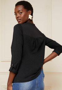 Haut noir à manches longues avec une texture douce, détail de pince au dos et poignets roulés. Associé à un jean en denim bleu clair.