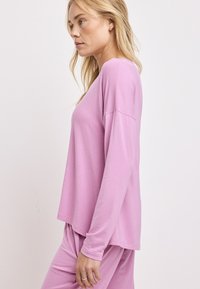 Długie rękawy, jasnoróżowa bluzka z miękkiego materiału, charakteryzująca się luźnym krojem i obniżonymi ramionami, stylizowana z dopasowanymi różowymi dołami.
