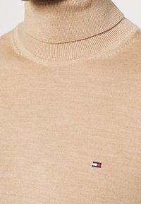 Pull à col roulé beige avec une texture côtelée et une coupe ajustée. Comprend un petit logo brodé en rouge, blanc et bleu sur la poitrine.