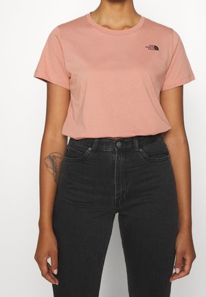 Persona que lleva una camiseta corta de manga corta rosa claro de The North Face y vaqueros negros de talle alto, mostrando un tatuaje de elefante en el brazo izquierdo.