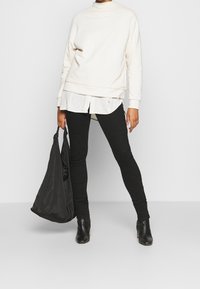 Sudadera crema con un cuello alto, superpuesta sobre una camisa blanca, combinada con pantalones ajustados negros y botas negras de tobillo, sosteniendo un bolso negro.