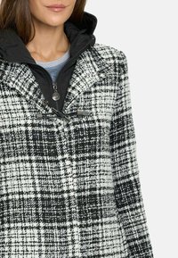 El abrigo de cuadros en blanco y negro presenta una tela texturizada con un alto cuello, cierre de cremallera y una capa interior negra. Los detalles incluyen herrajes de tipo toggle de imitación.