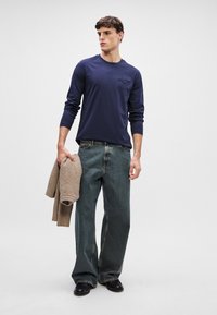 Mornarska majica s dolgimi rokavi in subtilnim logotipom, v kombinaciji s širokimi temnimi kavbojkami. Drži svetlo rjav pleten pulover. Črne čevlje.