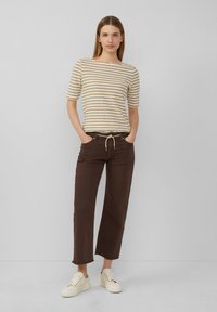 Pasiasty beżowo-kremowy t-shirt, w połączeniu z brązowymi spodniami zakończonymi w kostce i z regulowanym paskiem. Model nosi jasne trampki.