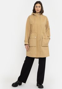 Khaki kapucňová parka s nastaviteľnou šnúrkou, dvoma prednými vreckami a uzávermi na cvoky. Má uvoľnený strih a hladký, odolný materiál.