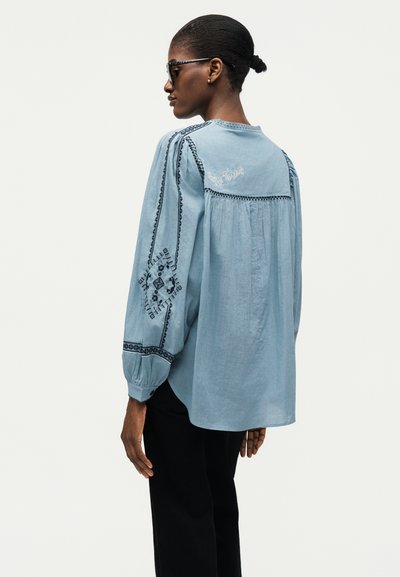 Γυναίκα που φοράει ανοιχτό μπλε μπλούζα με μαύρα κεντημένα σχέδια στα μανίκια και την πλάτη, μαύρο παντελόνι και σκούρα γυαλιά ηλίου, κοιτάζοντας πλάγια.