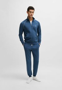 Tuta da palestra blu realizzata in tessuto liscio; giacca con zip e colletto alto con dettagli bianchi, abbinata a pantaloni aderenti con polsini elastici.