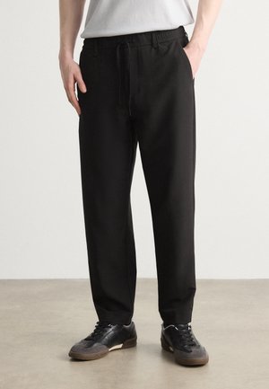 Mann trägt schwarze Freizeithose mit Kordelzug und schwarze Sneakers, steht auf neutralem Boden vor einer weißen Wand.