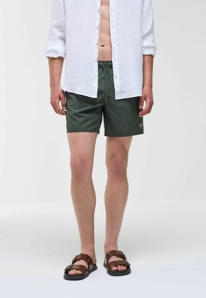 Uomo che indossa una camicia bianca sbottonata, pantaloncini da nuoto verde scuro con coulisse e sandali marroni a doppia fascia, in piedi davanti a uno sfondo uniforme.