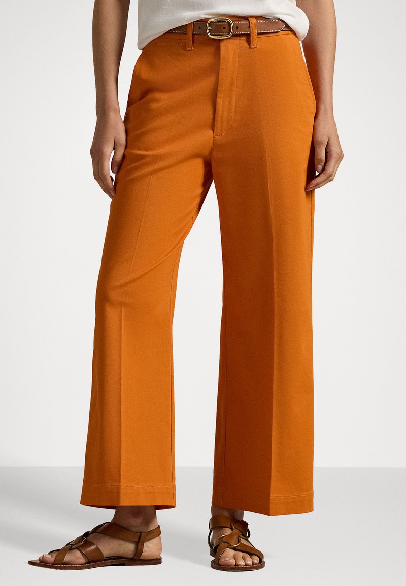Orange wide-leg trousers with a smooth texture and pleated front. Paired with brown strappy sandals and a brown belt with a square buckle.