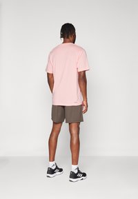 Camiseta de algodón rosa claro, de mangas cortas, combinada con pantalones cortos marrón oscuro. Zapatillas deportivas negras con detalles en blanco y logo, sobre un fondo neutro.
