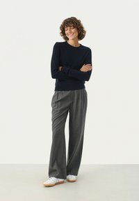 Navy Strickpullover, graue plissierte Hose und weiße Sneaker. Glatter Stoff, lässige Passform und minimalistisches Design ohne sichtbare Muster.