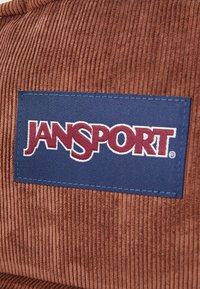 Καφέ πανί κορντούρας με μπλε ορθογώνια επιφάνεια που αναγράφει "JANSPORT" σε μπορντό γράμματα, περιγραμμένα με λευκή ραφή.